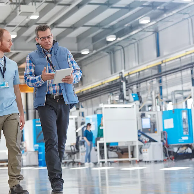 Two men walking through a customer site's production line. One holds a tablet, explaining to the other. Background shows the production/assembly line. Key visual for Expert Walk only.