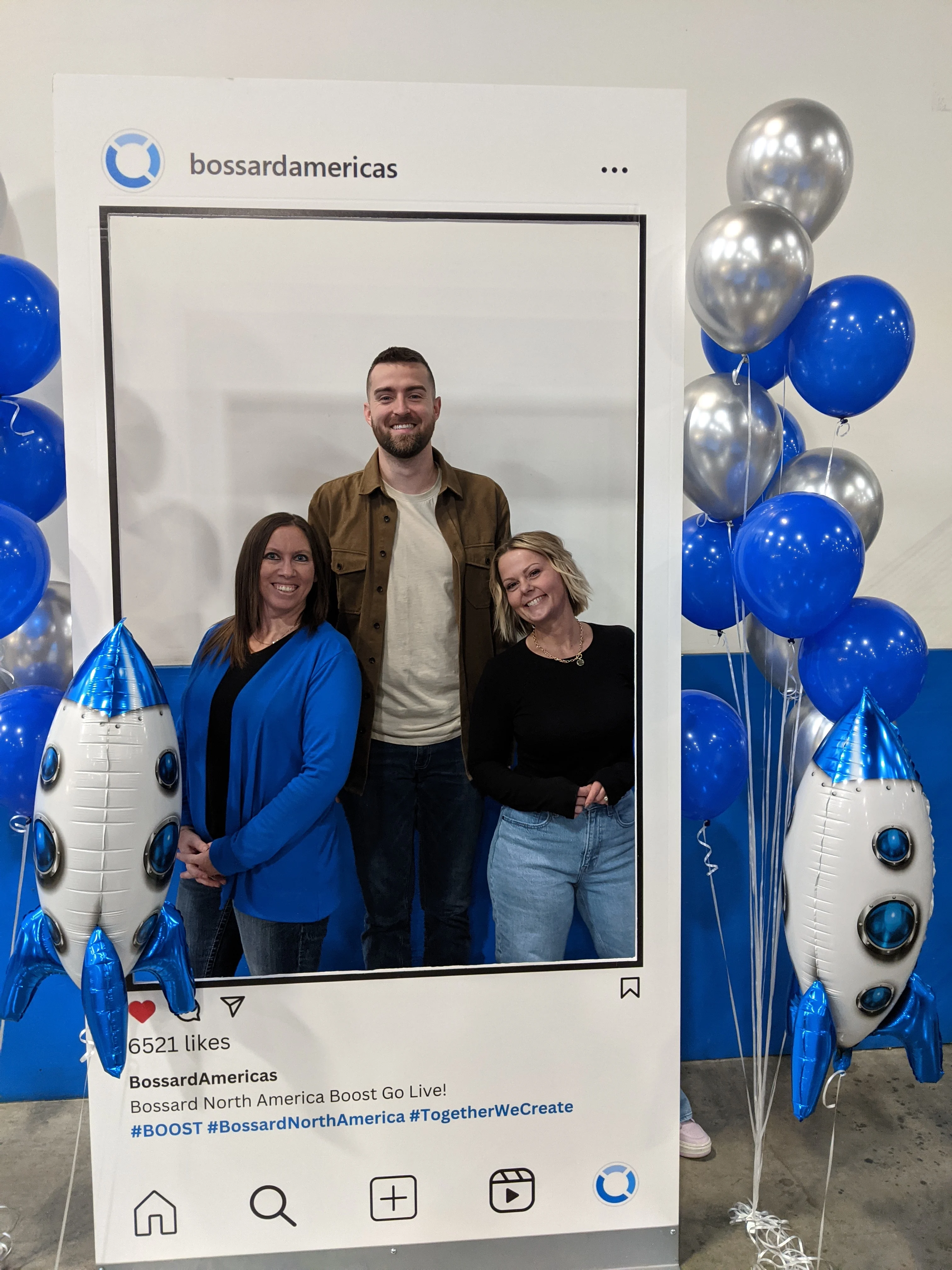 Three people pose together in a photo frame of a Bossard Americas social media post, surrounded by silver and blue balloons.