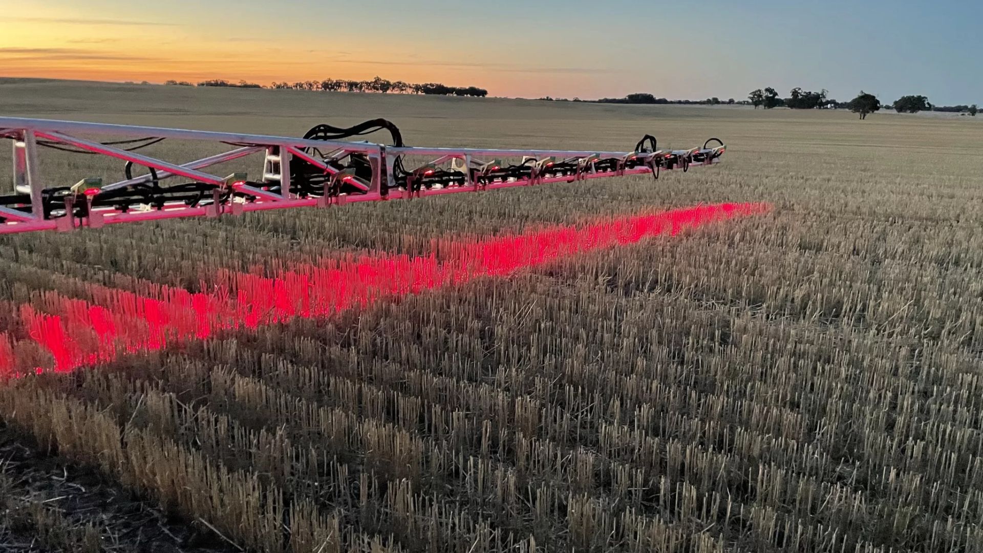 Red agricultural sprayer equipment casting bright red light across harvested stubble field at sunset with trees on horizon.