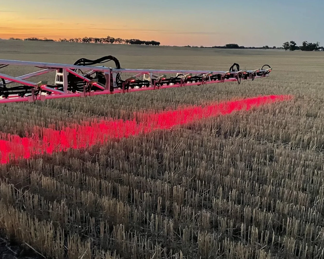 Red agricultural sprayer equipment casting bright red light across harvested stubble field at sunset with trees on horizon.