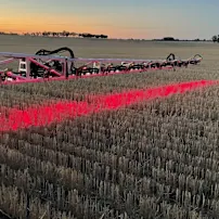 Red agricultural sprayer equipment casting bright red light across harvested stubble field at sunset with trees on horizon.
