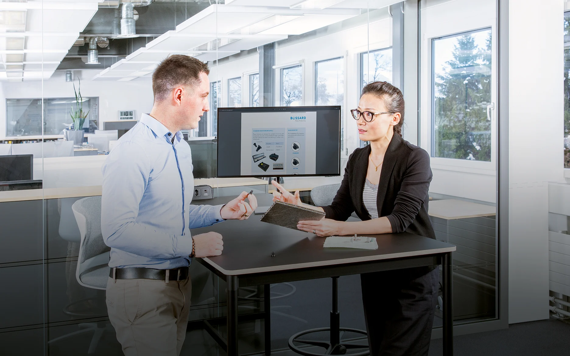 两个人在现代办公室的桌子上讨论复合材料的紧固问题;一个人拿着夹层板,另一个人拿着紧固件。