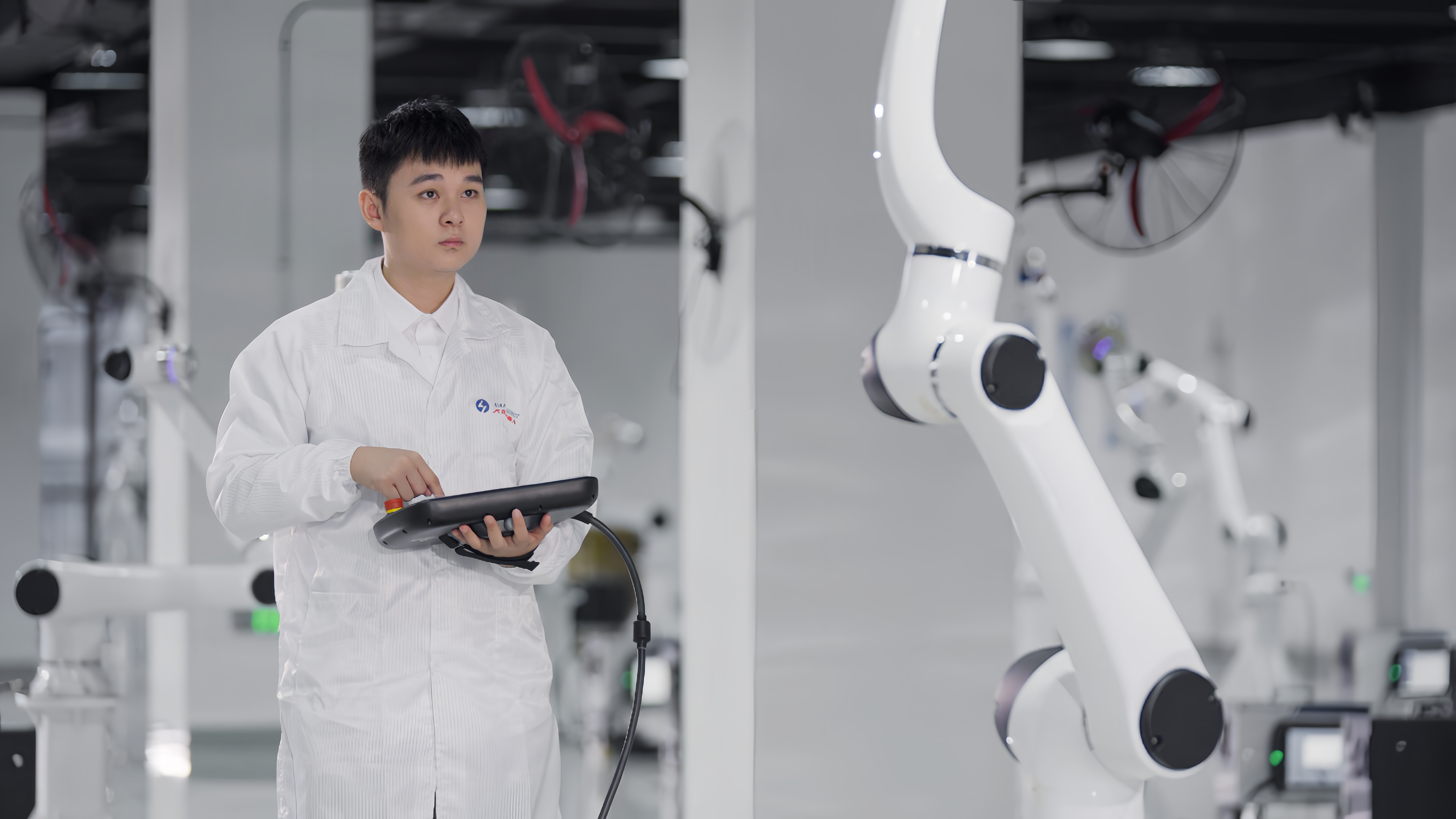 Technician in white lab coat holding control device stands next to white robotic arm in modern industrial facility.