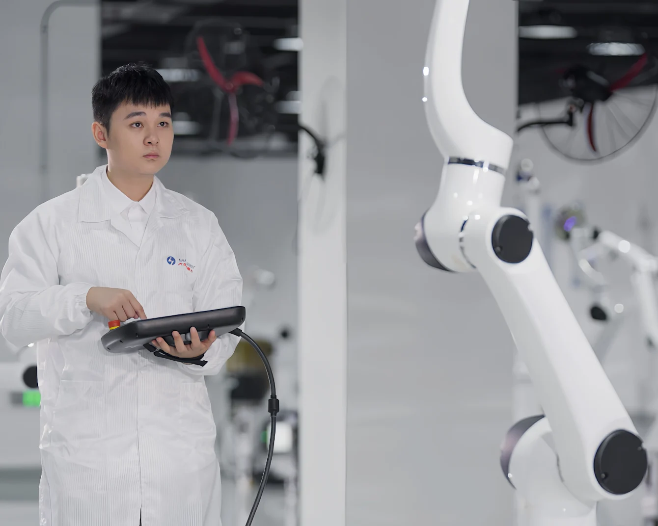 Technician in white lab coat holding control device stands next to white robotic arm in modern industrial facility.