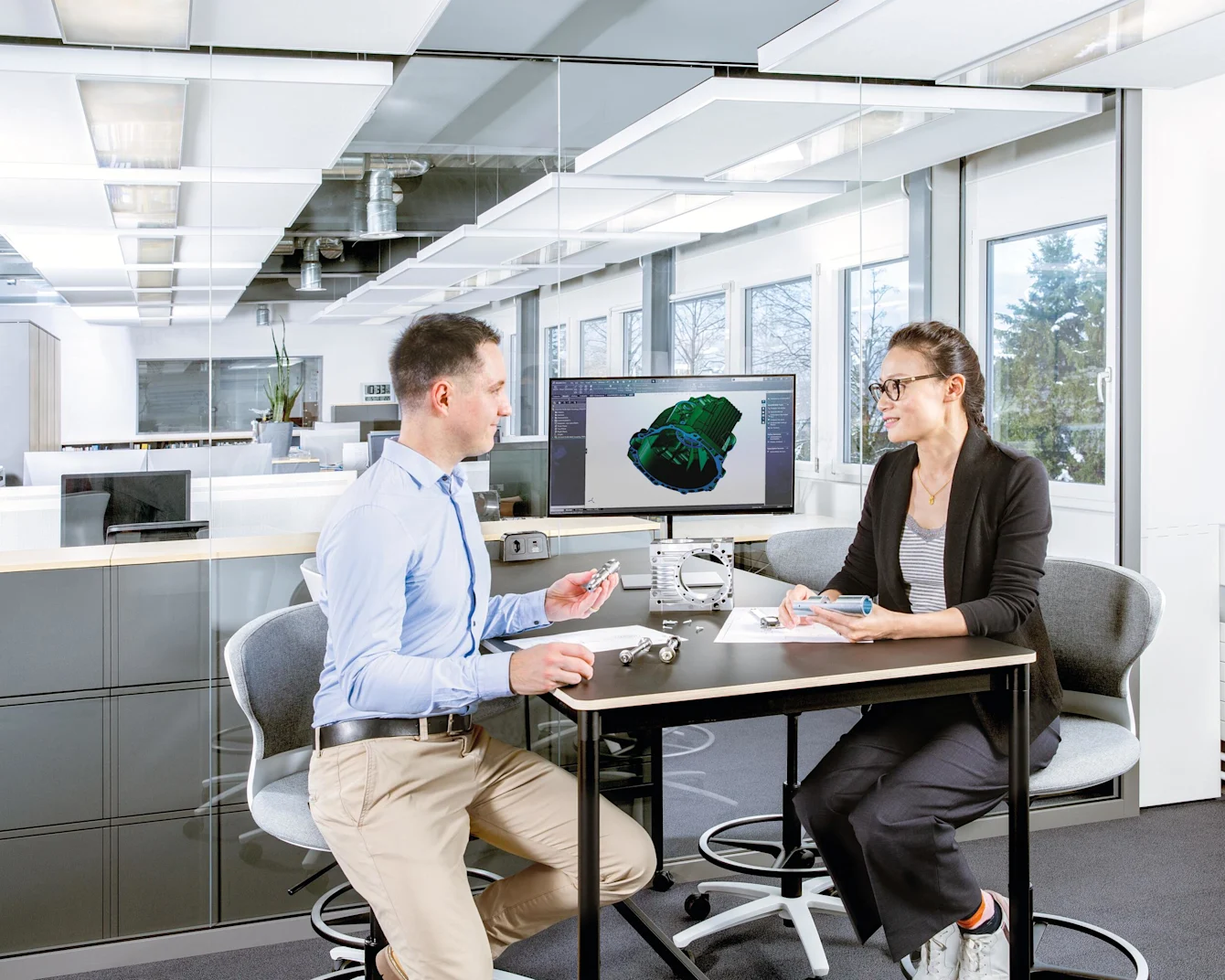 A male and female sit at a table with a screen displaying a technical drawing. On the table are a physical application, a technical drawing, and several screws. Key visual for Expert Design only.