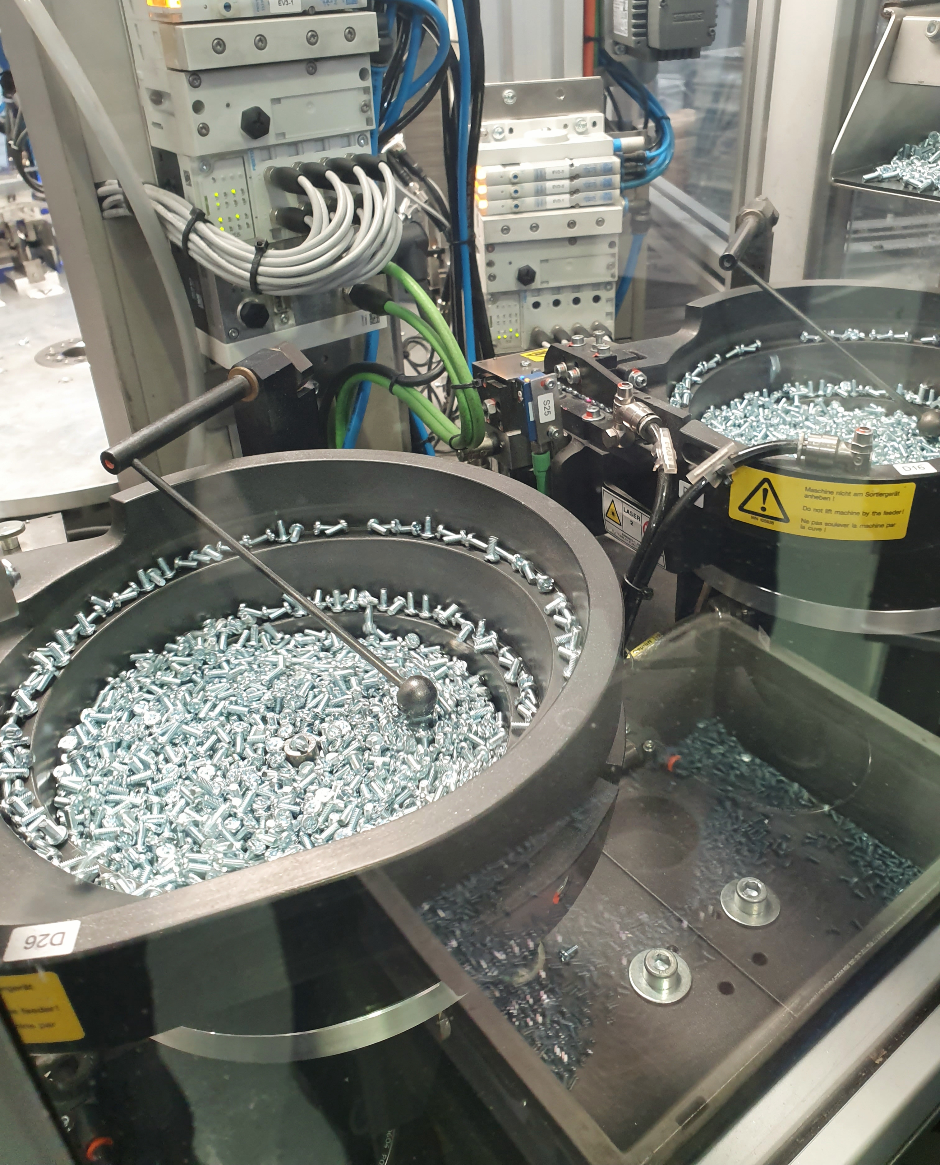 Industrial machine with two circular trays filled with silver screws, surrounded by cables and control panels.