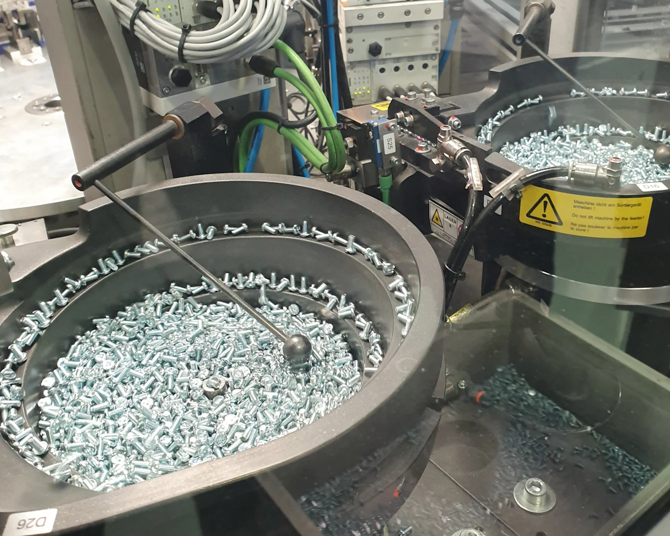 Industrial machine with two circular trays filled with silver screws, surrounded by cables and control panels.