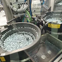 Industrial machine with two circular trays filled with silver screws, surrounded by cables and control panels.