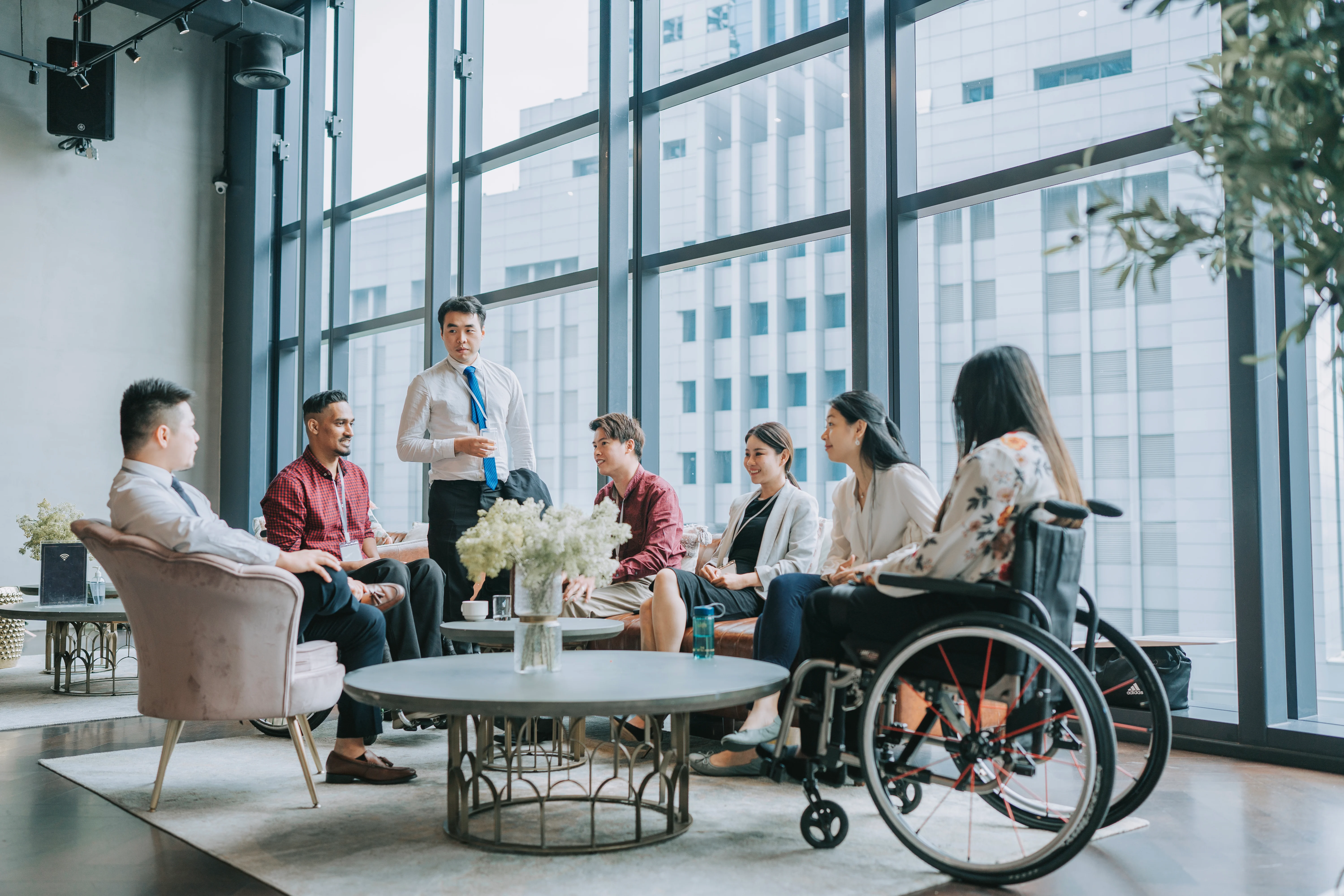 A group of individuals in a lobby, with one person in a wheelchair and another standing nearby, engaged in conversation.