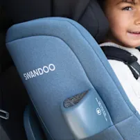 Child in a blue Swandoo car seat, smiling, wearing a white shirt and jeans, seated in a car's backseat.