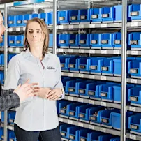 A man and a woman standing in front of a rack of blue Bossard Smartbins at Weibel