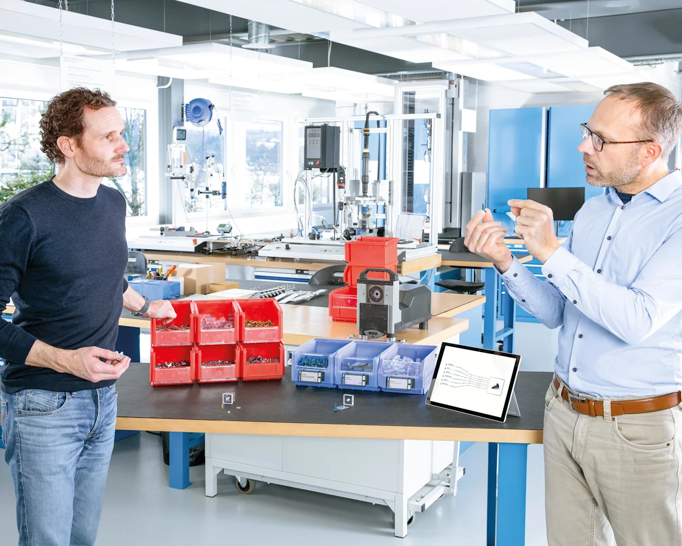 Engineer explains the benefits of multifunctional screws to a customer at a table with red and blue boxes of fasteners. A tablet is showing the potential of streamlining the number of C-parts. Key visual for Expert Assortment Analysis only.