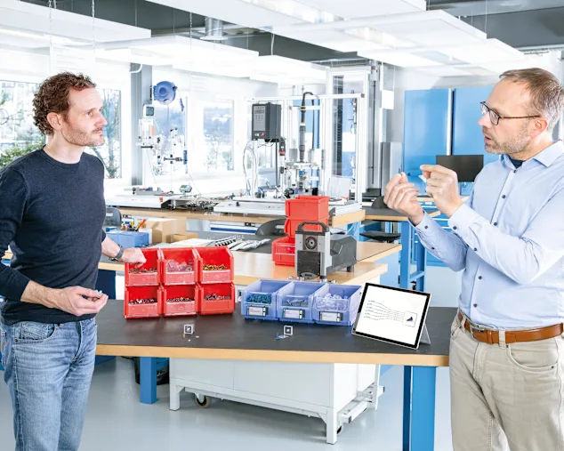 Engineer explains the benefits of multifunctional screws to a customer at a table with red and blue boxes of fasteners. A tablet is showing the potential of streamlining the number of C-parts. Key visual for Expert Assortment Analysis only.