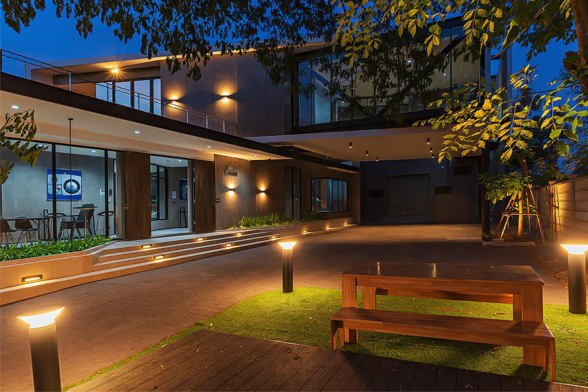 Modern house exterior at dusk with warm lighting, large windows, and a wooden bench on a grassy area, surrounded by trees.