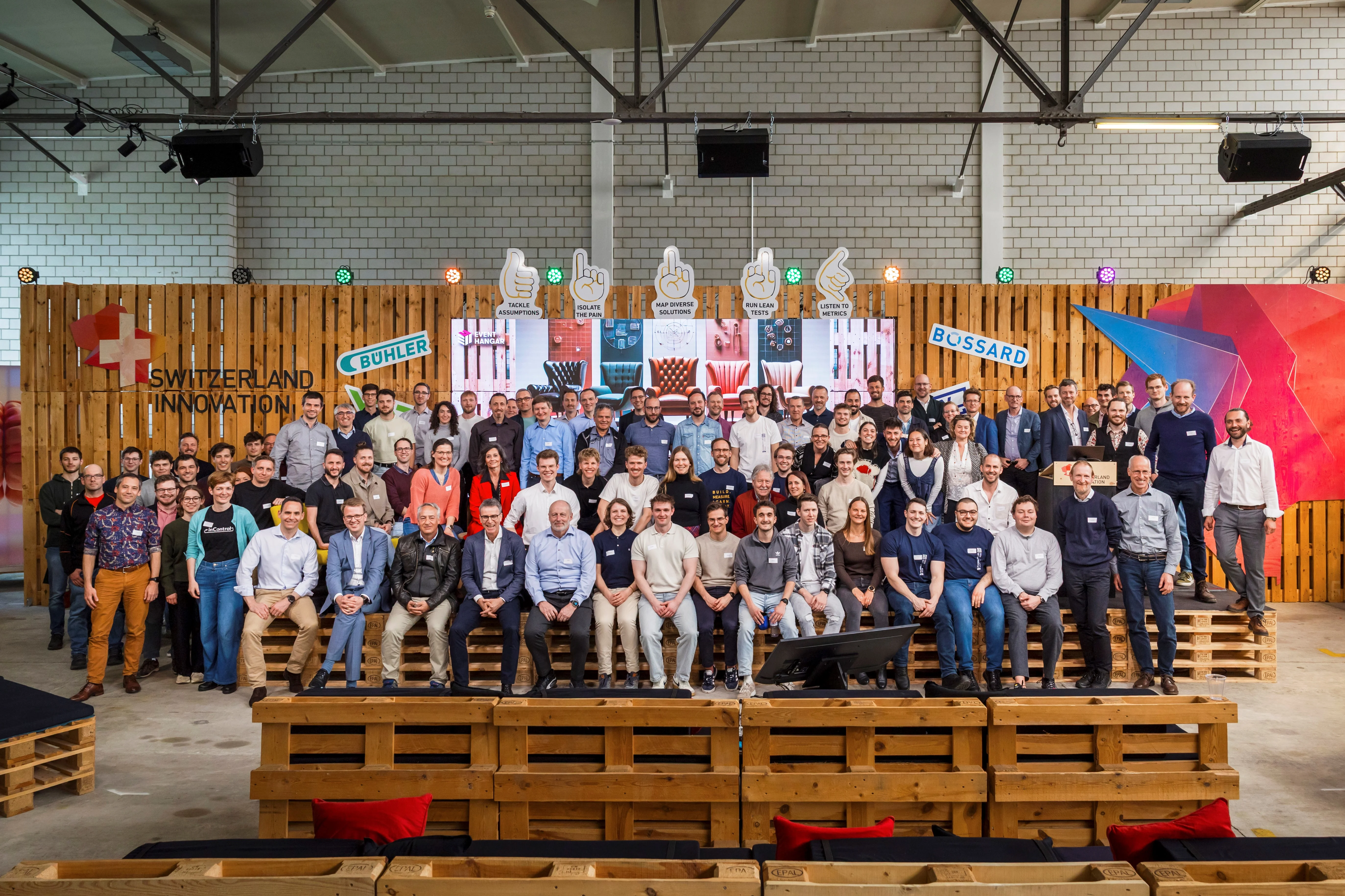 A large group of people pose on wooden benches in a warehouse setting, with "Switzerland Innovation" and logos in the background.