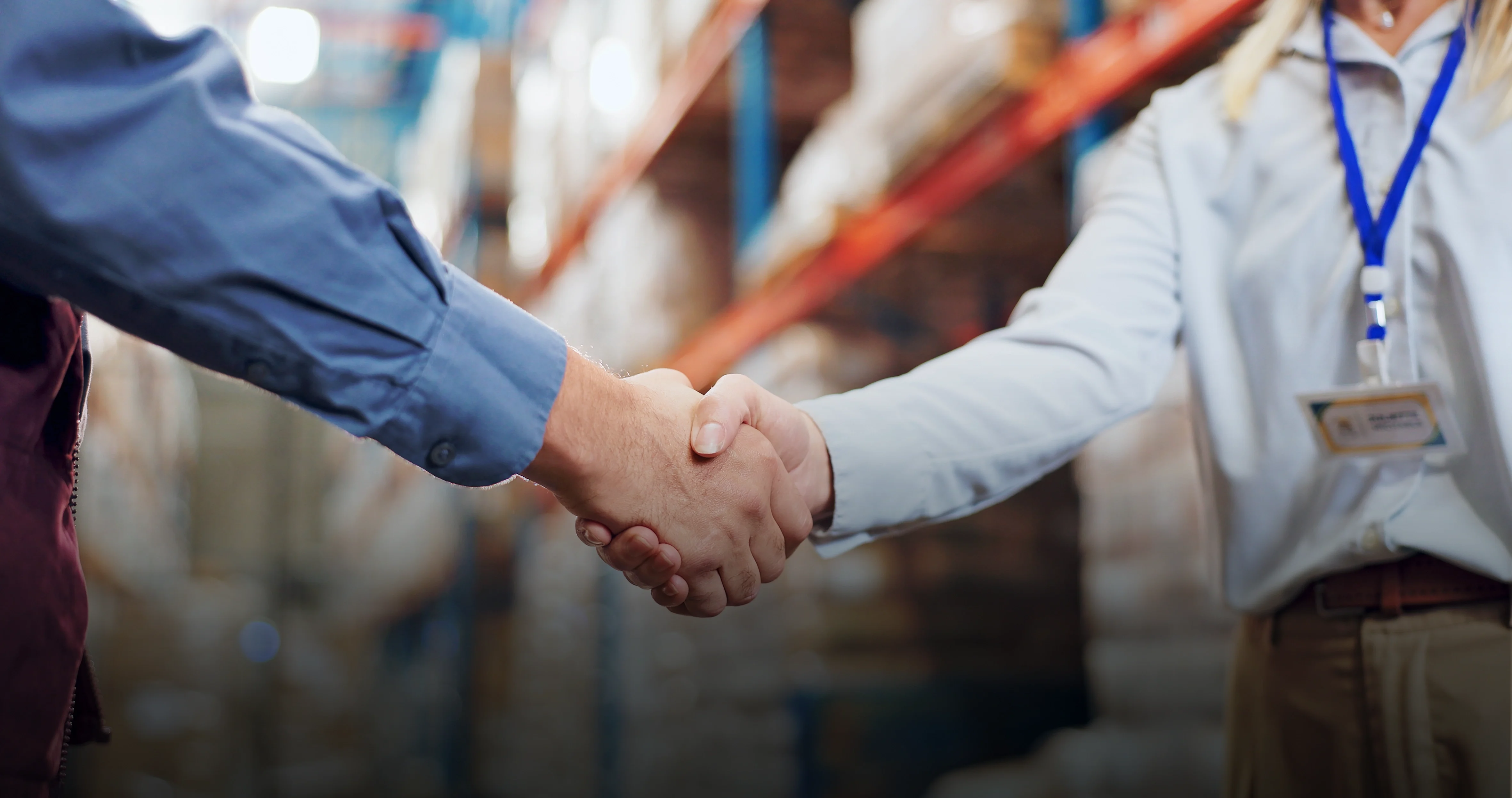 Two people shaking hands in a warehouse, one wearing a blue shirt, the other with a name badge, shelves in the background.