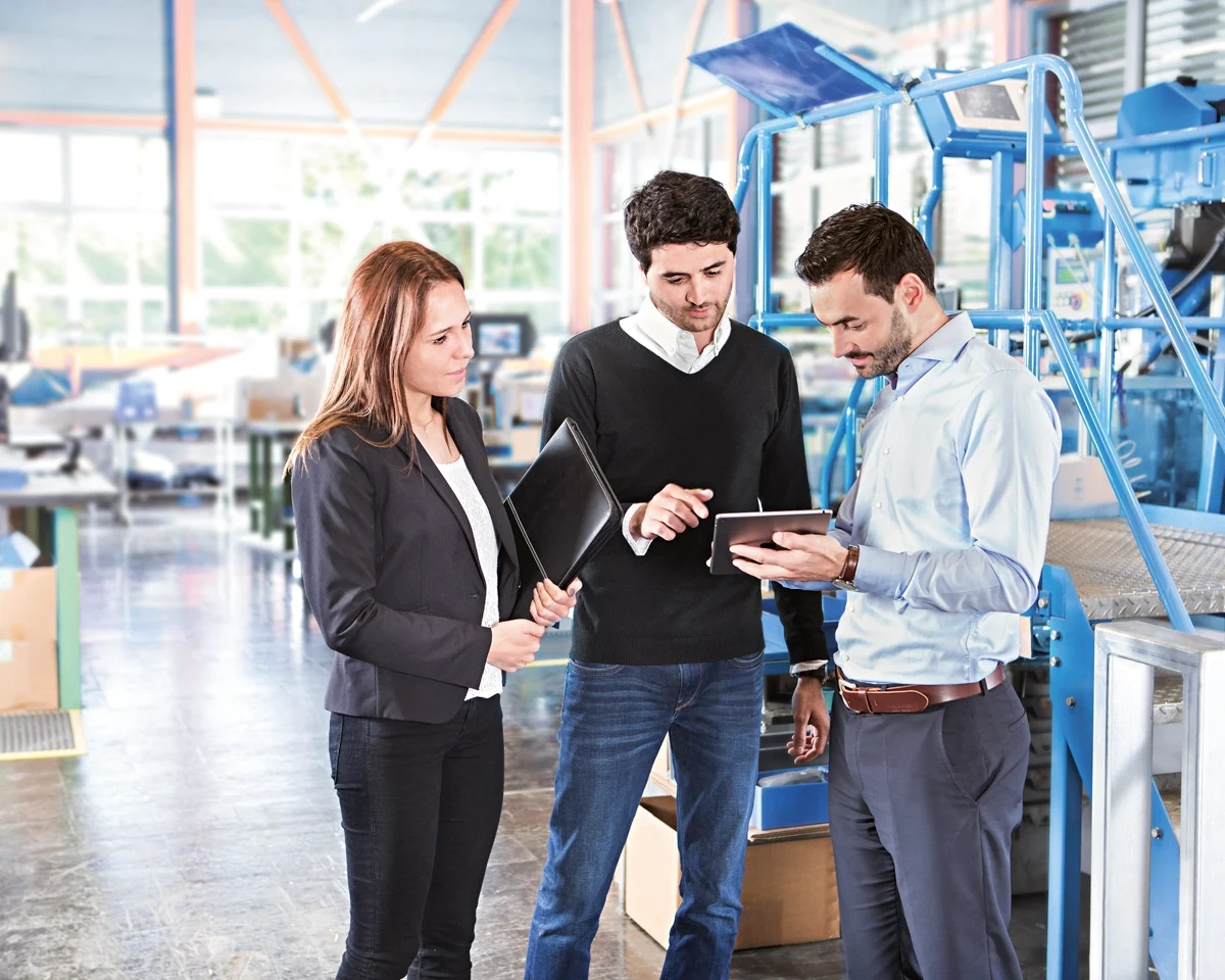 Three individuals in a factory observe a tablet, engaged in discussion about their work or project.