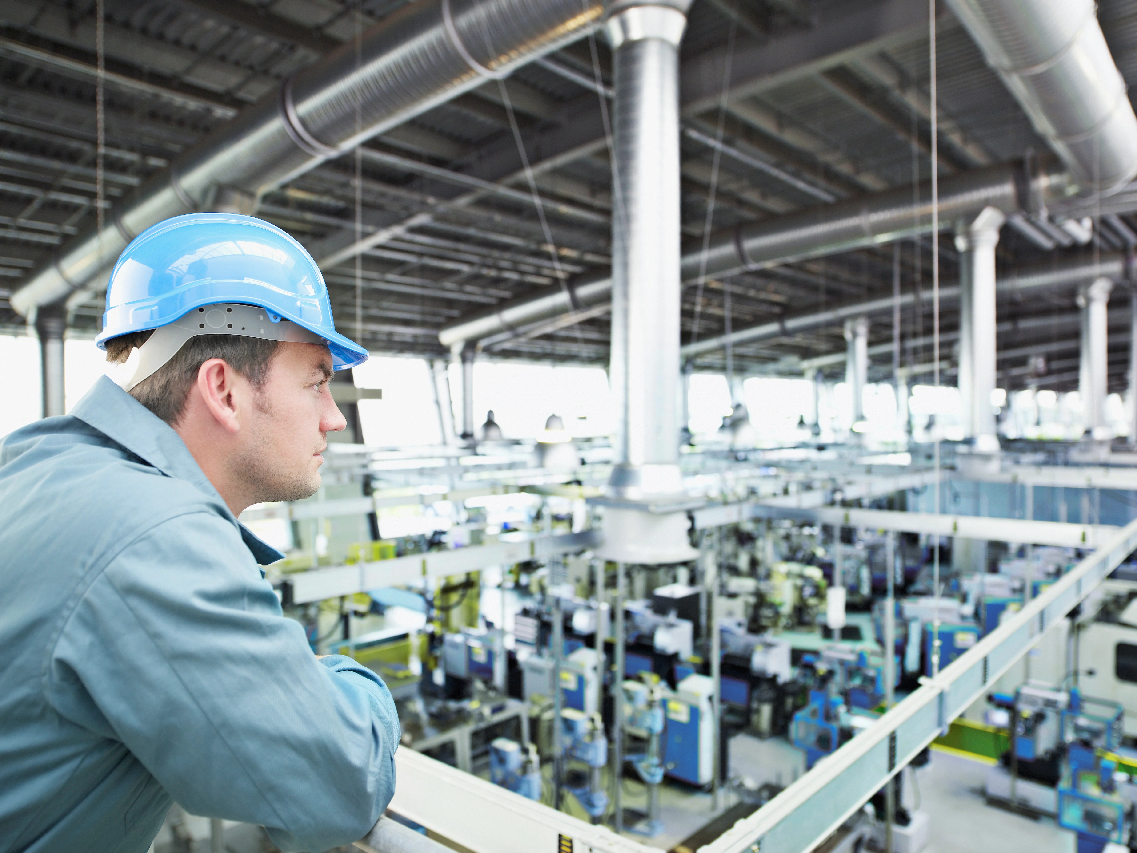 A factory worker wearing a hard hat observes machinery in a factory setting, ensuring safety and operational efficiency.
