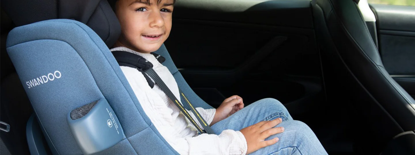 Child in a blue Swandoo car seat, smiling, wearing a white shirt and jeans, seated in a car's backseat.