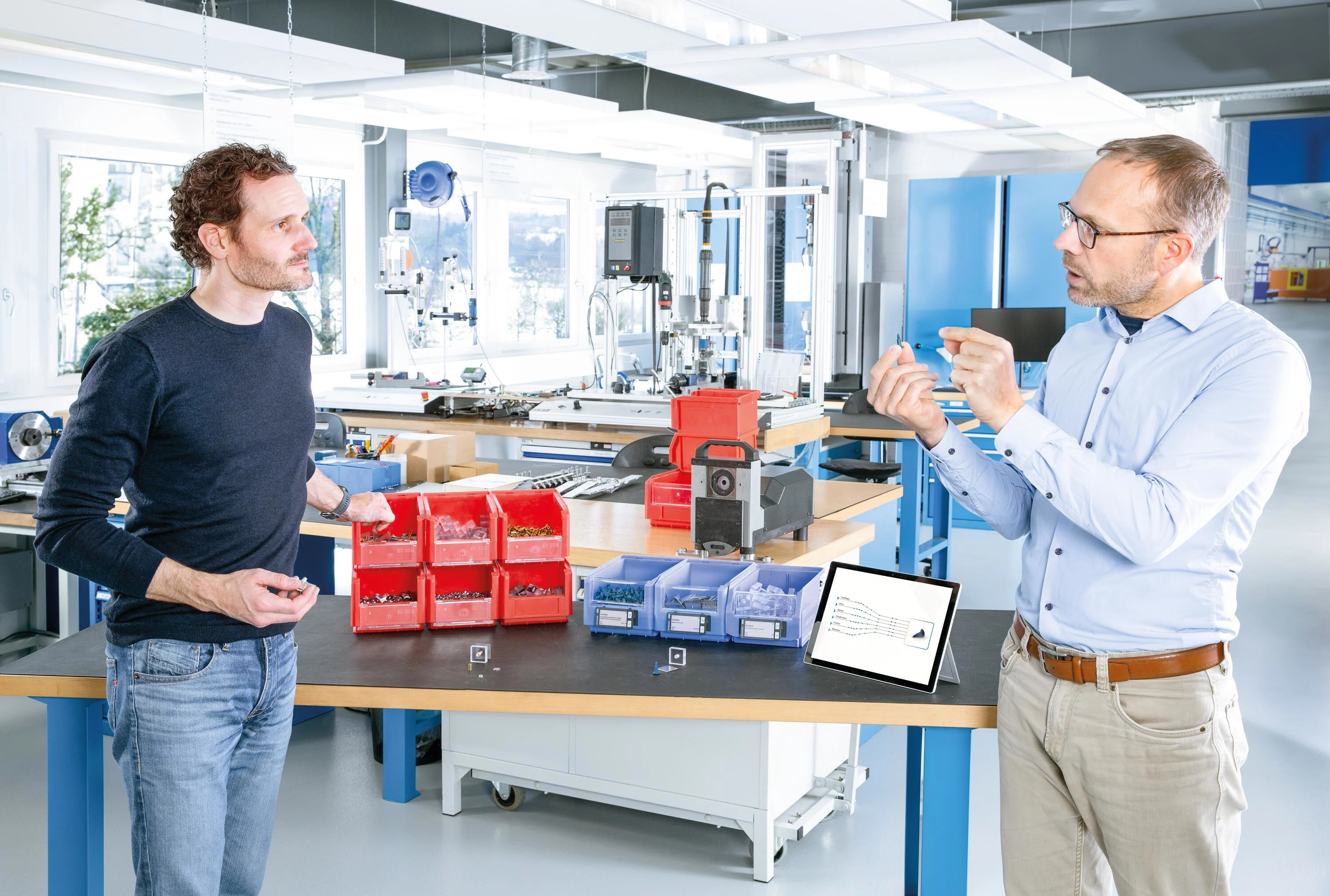 Engineer explains the benefits of multifunctional screws to a customer at a table with red and blue boxes of fasteners. A tablet is showing the potential of streamlining the number of C-parts. Key visual for Expert Assortment Analysis only.